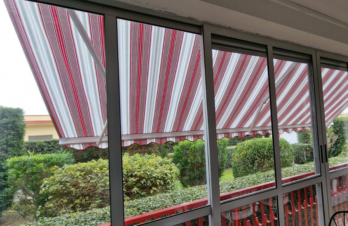 Tenda a bracci estensibili con telo a righe rosse e bianche vista da una veranda vetrata, siepi verdi in giardino
