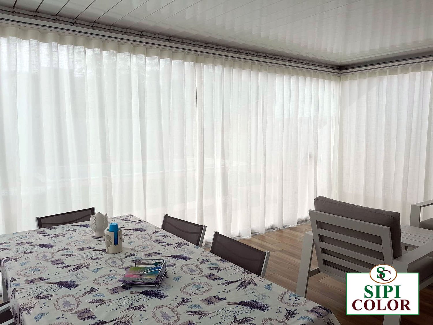Tende bianche velate su binario chiudono una veranda con tavolo, tovaglia a lavanda e sedie grigie, posa Sipi Color